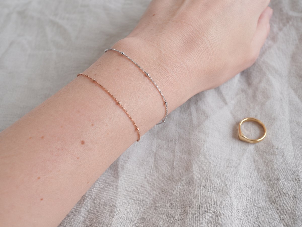 Muñeca con dos pulseras finas de plata y oro rosa, con un pequeño anillo dorado sobre una tela beis