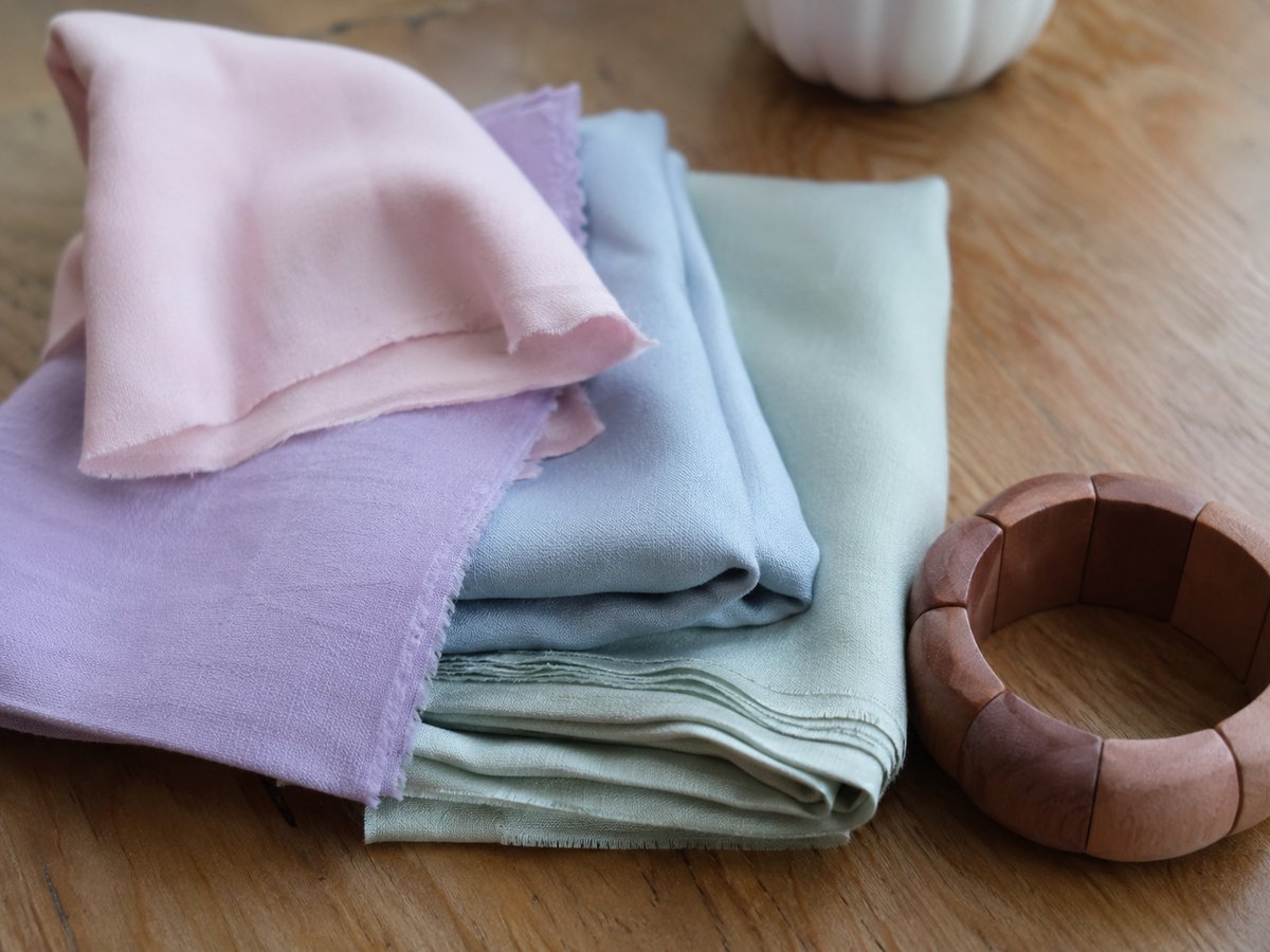 Cuadrados de tela en tonos pastel rosa, lila, azul cielo y verde salvia sobre una mesa de madera con una pulsera de madera